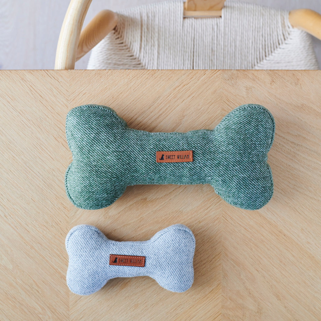 Large green and small grey tweed dog bone toys side by side, showing the two sizes on a wooden table