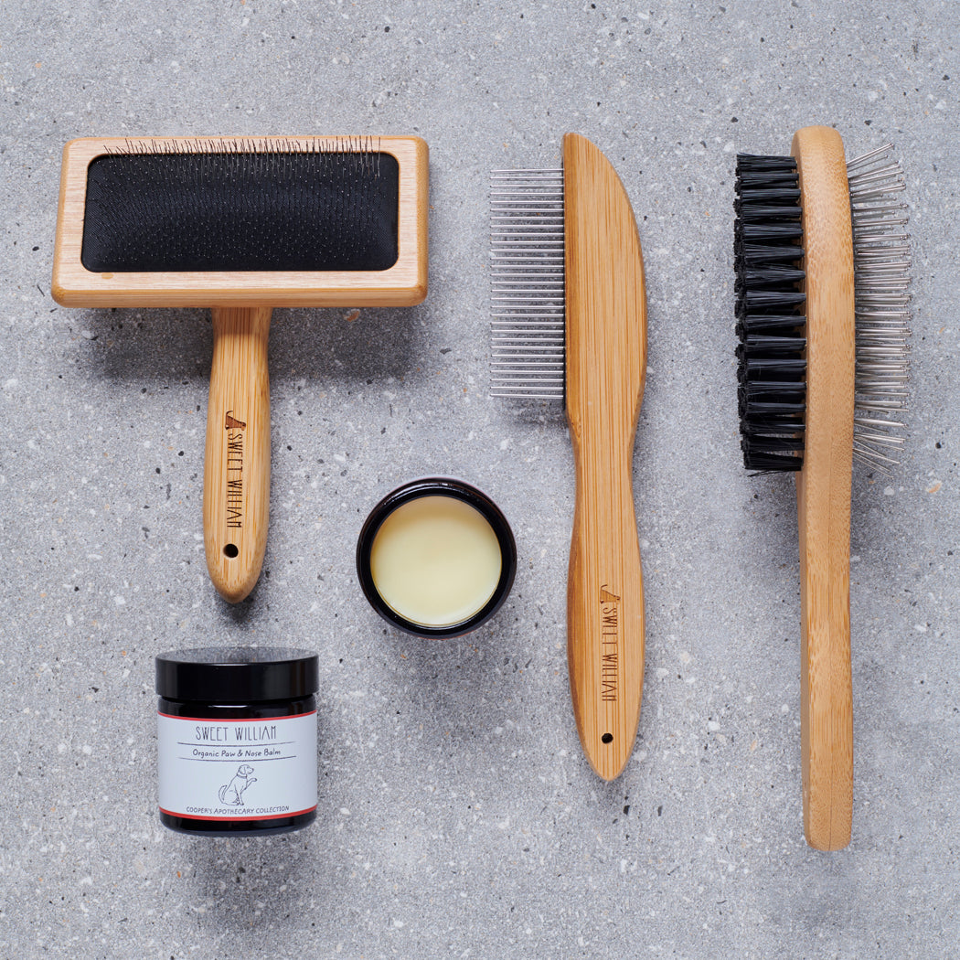 Sweet William bamboo dog grooming tools including — slicker brush, metal comb, double sided brush and Organic Paw & Nose Balm on grey stone surface, dog grooming gift set