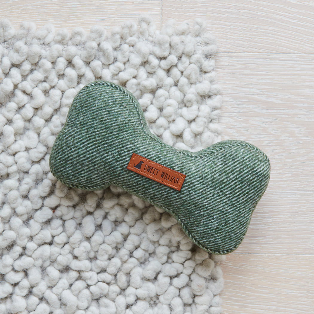Green tweed dog bone toy on a chunky white rug, showing its woven fabric and Sweet William label