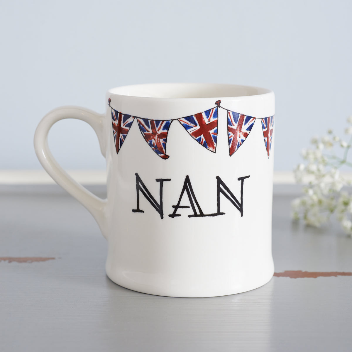 Ceramic Nan mug with hand-drawn Union Jack bunting, styled beside delicate white flowers, a thoughtful gift for a much-loved grandparent