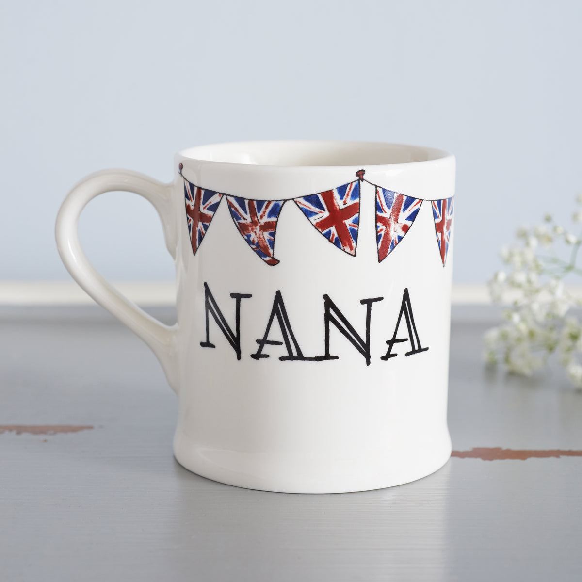 Ceramic Nana mug with hand-drawn Union Jack bunting, styled beside delicate white flowers, a thoughtful gift for a much-loved grandparent
