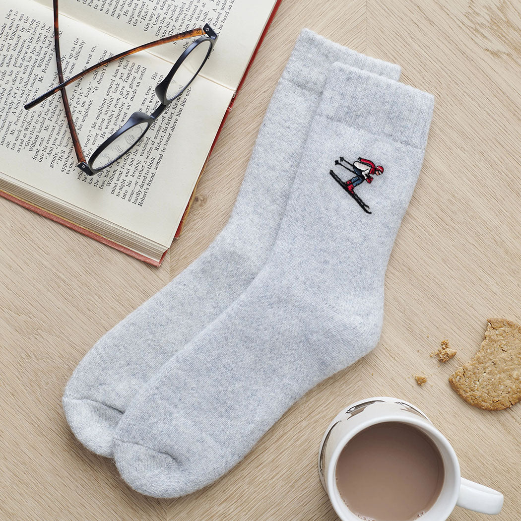 Embroidered skier wool socks laid flat beside a book, glasses and a cup of tea — cosy ski lover gift by Sweet William Designs