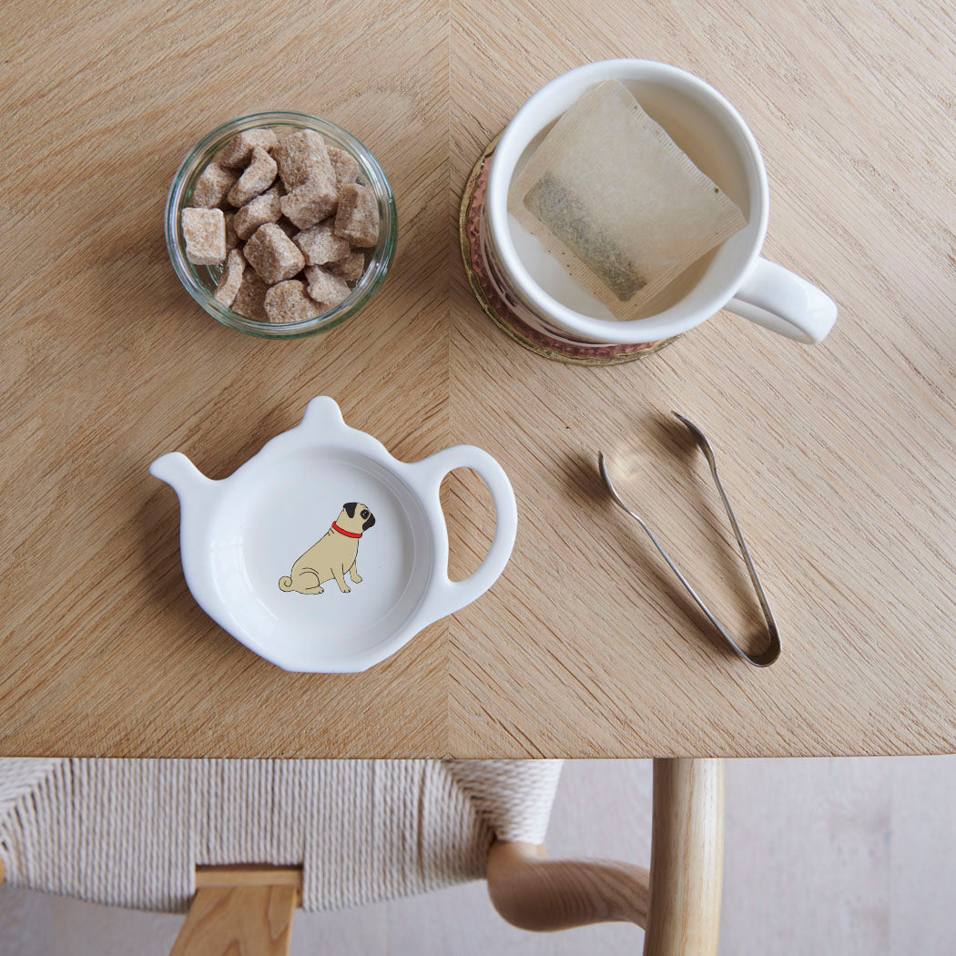White teapot-shaped Pug teabag dish with a fawn Pug illustration and red collar, beside a mug, sugar cubes and sugar tongs