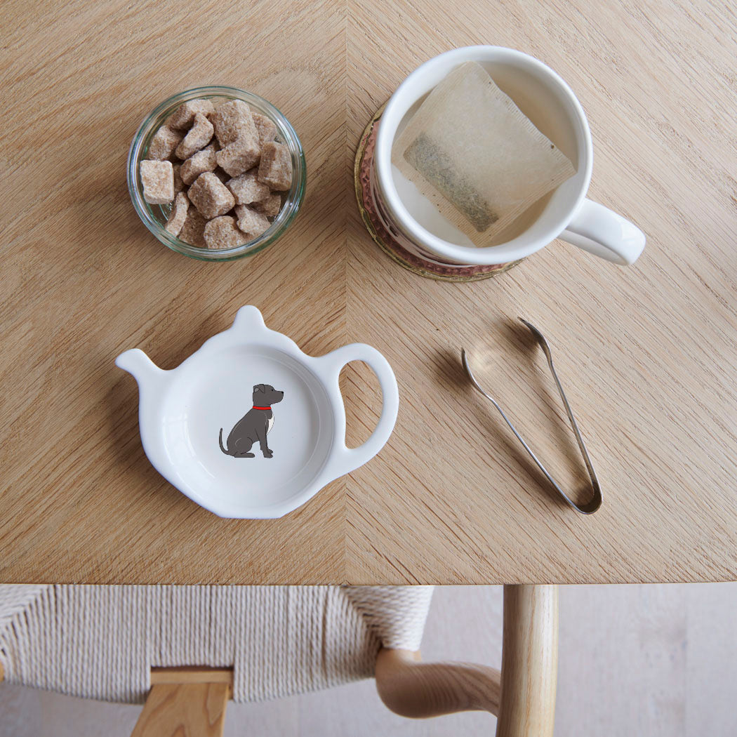 White teapot-shaped Staffie teabag dish with a grey dog illustration, white chest and red collar, beside a mug, sugar cubes and sugar tongs