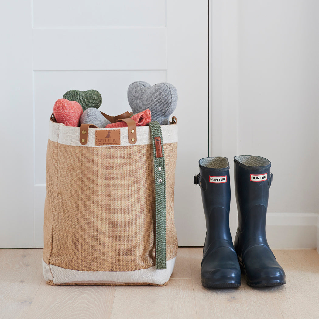 Grey, green and orange tweed dog bone toys peeking out of a Sweet William jute bag beside a pair of Hunter wellies