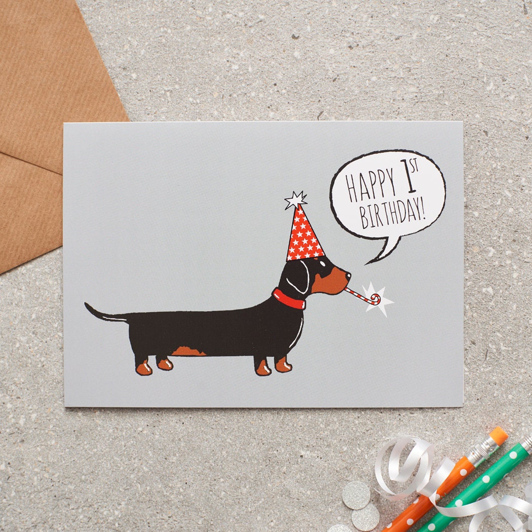 Dachshund 1st birthday card featuring a black and tan sausage dog in a red party hat with a “Happy 1st Birthday!” message, designed by Sweet William Designs