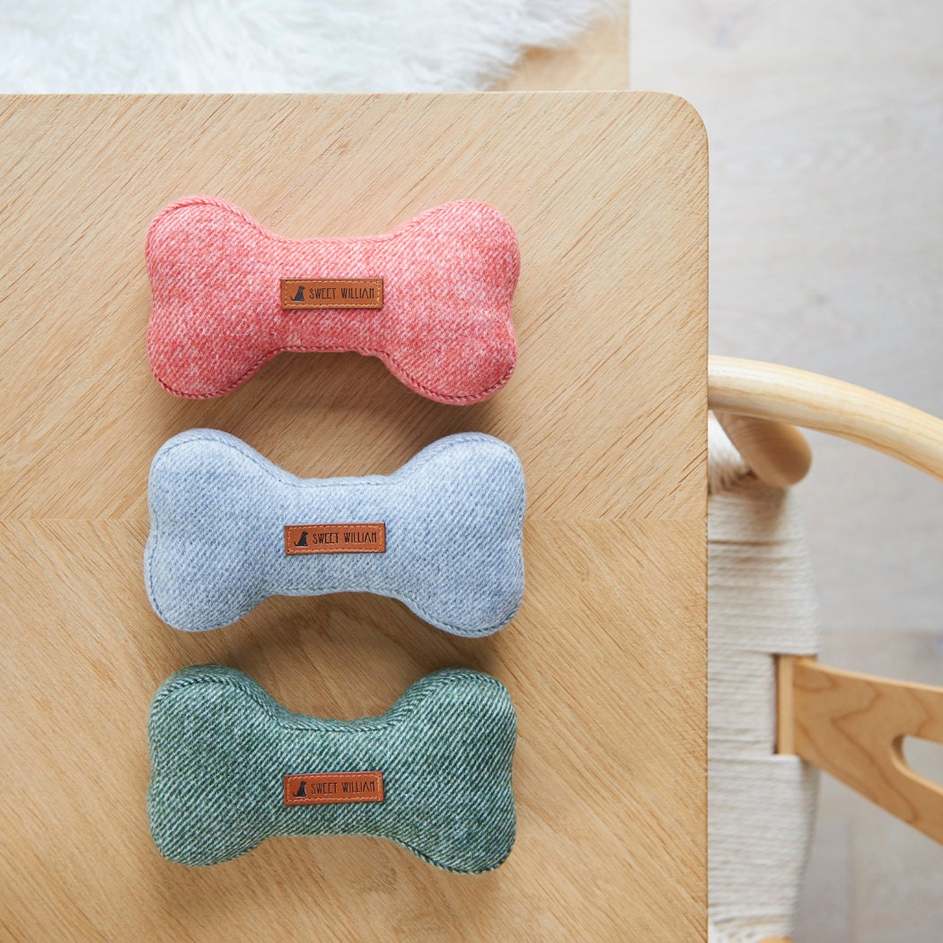 The full Sweet William tweed dog bone toy colour range in orange, grey and green, arranged together on a wooden table