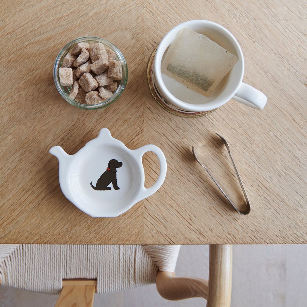 White teapot-shaped Chocolate Cocker Spaniel teabag dish with a brown dog illustration and red collar, beside a mug, sugar cubes and sugar tongs