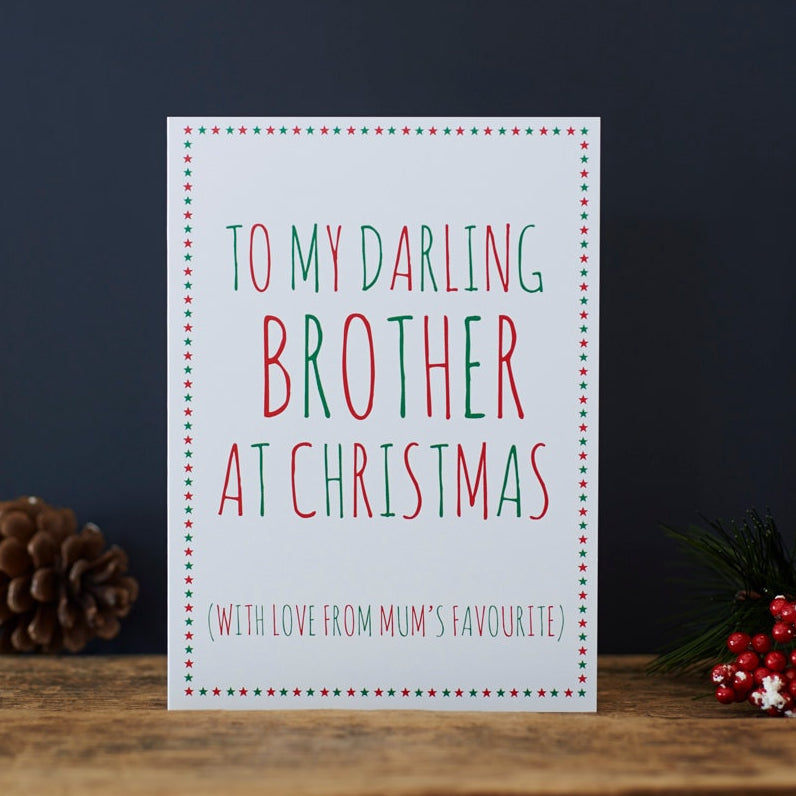 White Brother Christmas card with red and green lettering reading “To My Darling Brother at Christmas” and “With love from Mum’s favourite”, a star border, pine cone and red berries beside it.