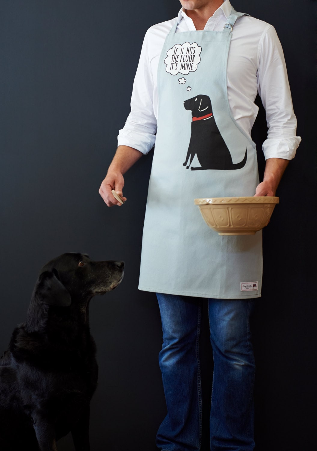 Person wearing Black Labrador 'If It Hits The Floor It's Mine' apron holding a mixing bowl, with William the Black Lab looking up hopefully — organic cotton dog lover gift by Sweet William Designs