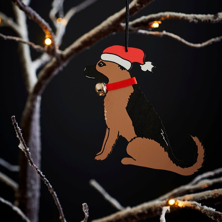 German Shepherd Christmas tree decoration hanging on lit winter tree — wooden dog decoration with Father Christmas hat and jingle bell collar by Sweet William Designs