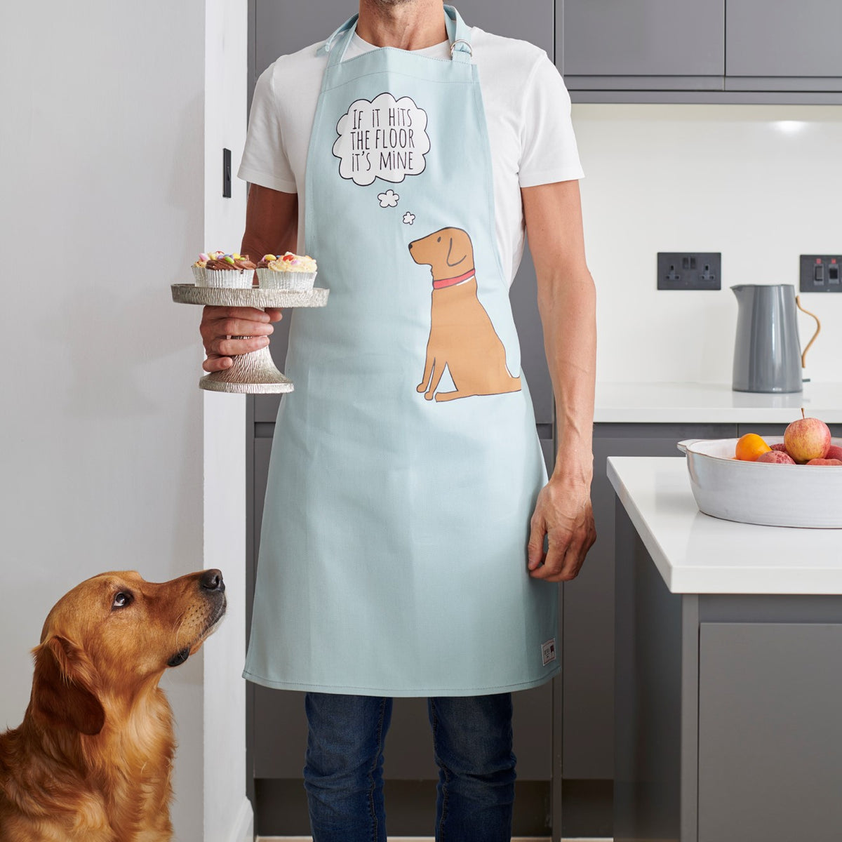 Person wearing a Fox Red Labrador “If It Hits The Floor It’s Mine” apron, holding a cake stand while Cooper looks up hopefully — organic cotton dog lover gift by Sweet William Designs
