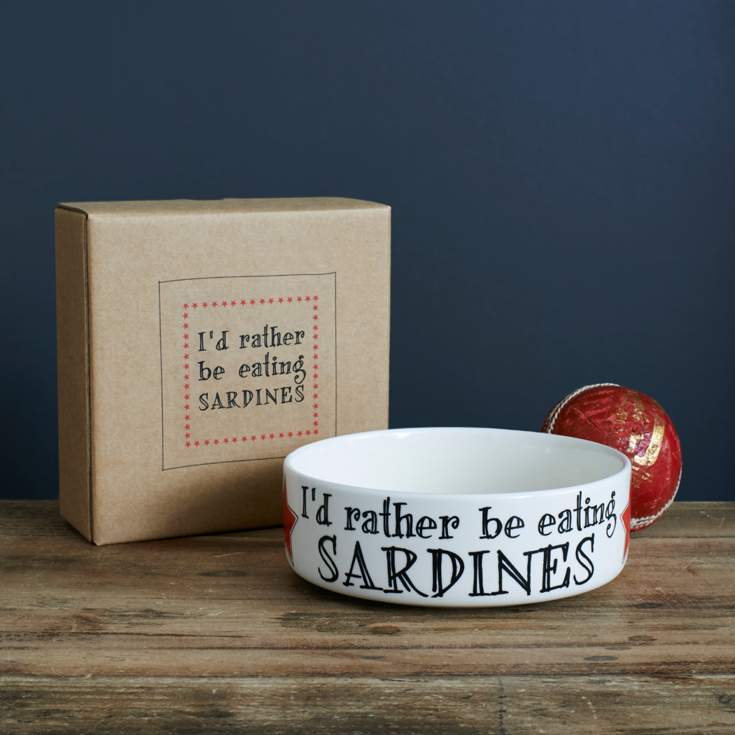 White porcelain “I’d Rather Be Eating Sardines” cat bowl with a kraft gift box and red ball on a wooden table