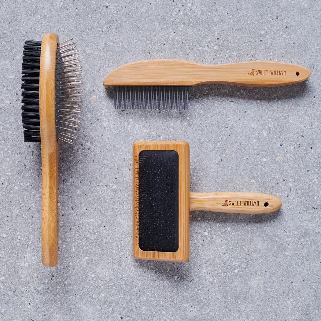 Sweet William bamboo dog grooming collection including — metal comb, slicker brush and double sided brush on grey stone surface, dog grooming gift set