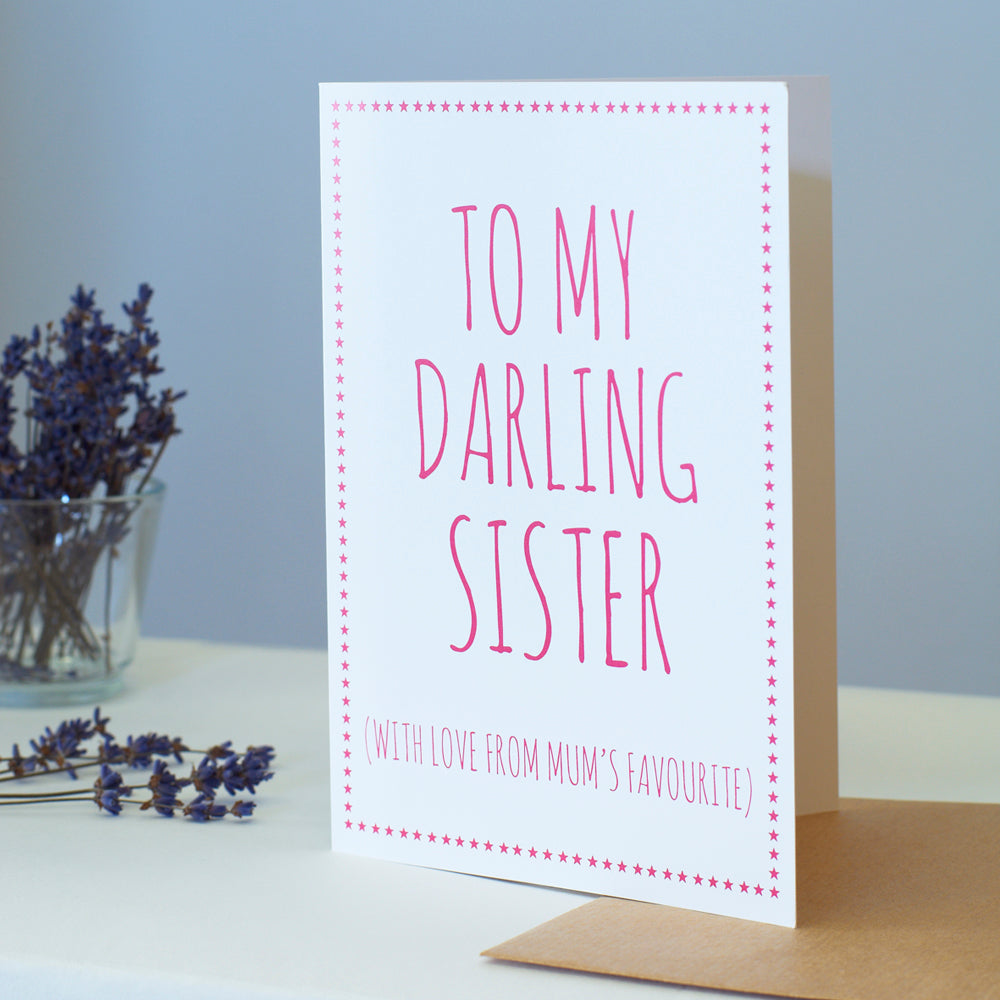White Sister card with pink lettering reading “To My Darling Sister” and “With love from Mum’s favourite”, a pink star border, kraft envelope and dried flowers beside it.