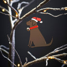 Chocolate  Labrador Christmas tree decoration hanging on lit winter tree — wooden dog decoration with Father Christmas hat and jingle bell collar by Sweet William Designs
