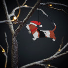 Basset Hound Christmas tree decoration hanging on lit winter tree — wooden dog decoration with Father Christmas hat and jingle bell collar by Sweet William Designs