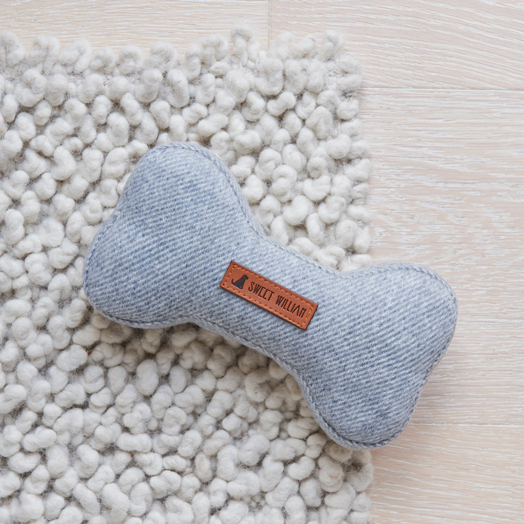 Close-up of the grey tweed dog bone toy on a soft white rug, showing its woven fabric and Sweet William label