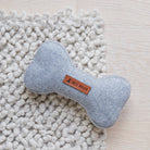 Close-up of the grey tweed dog bone toy on a soft white rug, showing its woven fabric and Sweet William label