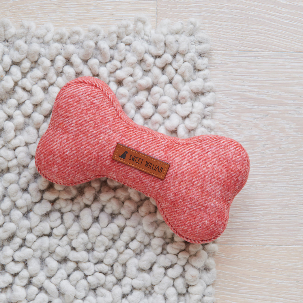Orange tweed dog bone toy resting on a chunky white rug, showing its woven fabric and Sweet William label