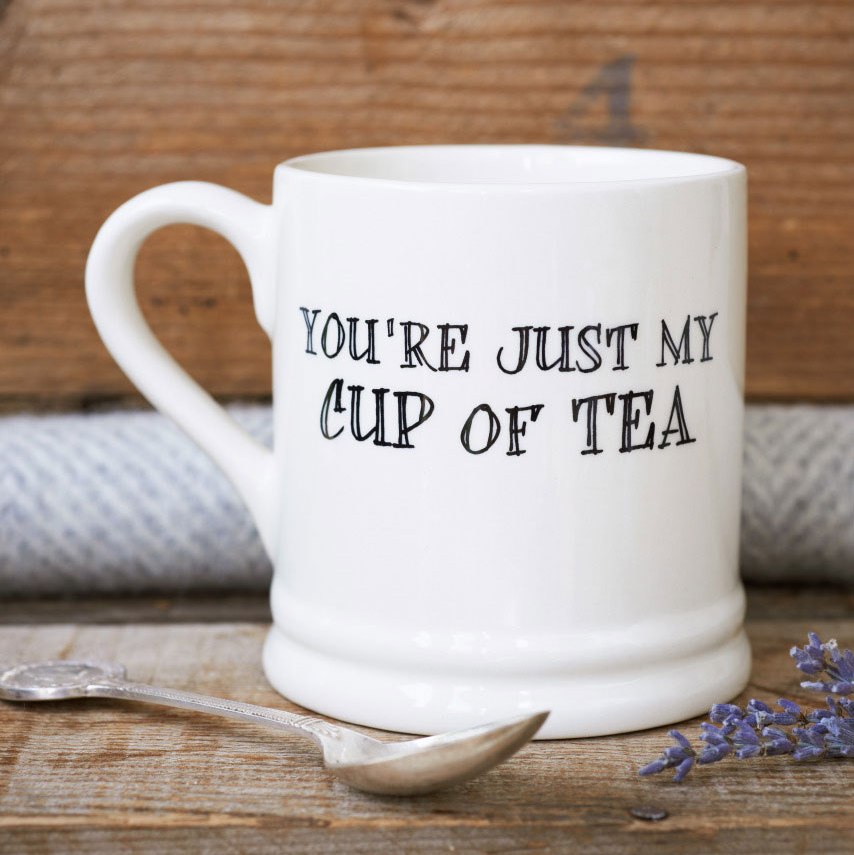 Porcelain mug with “You’re just my cup of tea” lettering, a silver spoon and sprigs of lavender on a rustic wooden table