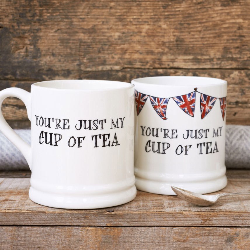 Two porcelain mugs with “You’re just my cup of tea” lettering, one plain and one decorated with Union Jack bunting, beside a silver spoon