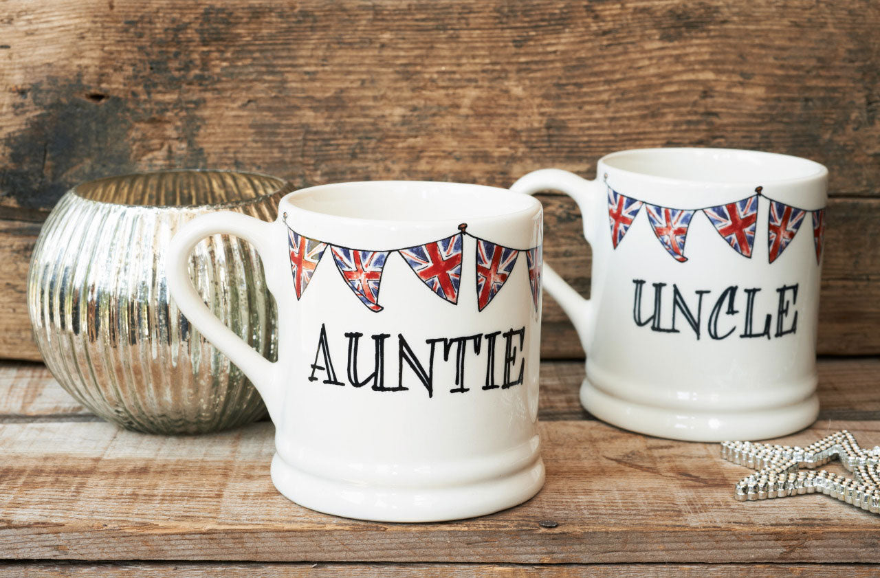 Ceramic Auntie and Uncle mugs from Sweet William Designs with Union Jack bunting, styled with a silver candle and star decoration on a rustic wooden surface