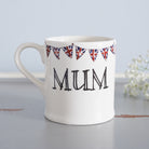 Ceramic Mum mug with hand-drawn Union Jack bunting, styled beside delicate white flowers as a thoughtful gift for a much-loved Mum