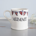 Ceramic Mummy mug decorated with hand-drawn Union Jack bunting, shown beside delicate white flowers