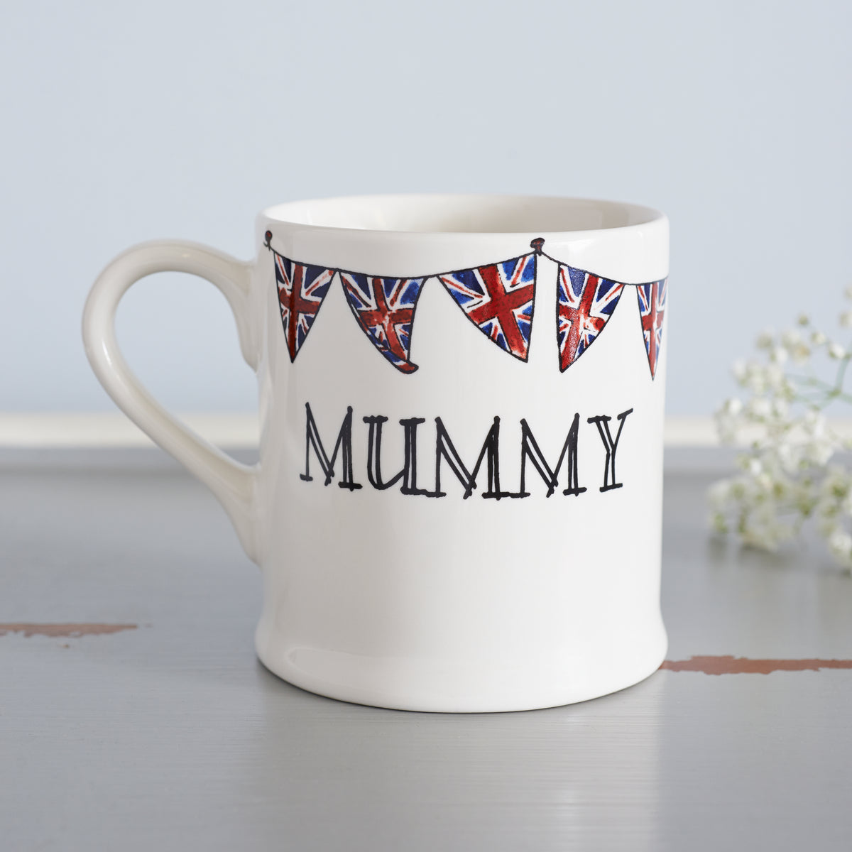 Ceramic Mummy mug decorated with hand-drawn Union Jack bunting, shown beside delicate white flowers