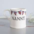 Ceramic Nanny mug with hand-drawn Union Jack bunting, styled beside delicate white flowers, a thoughtful gift for a much-loved grandparent