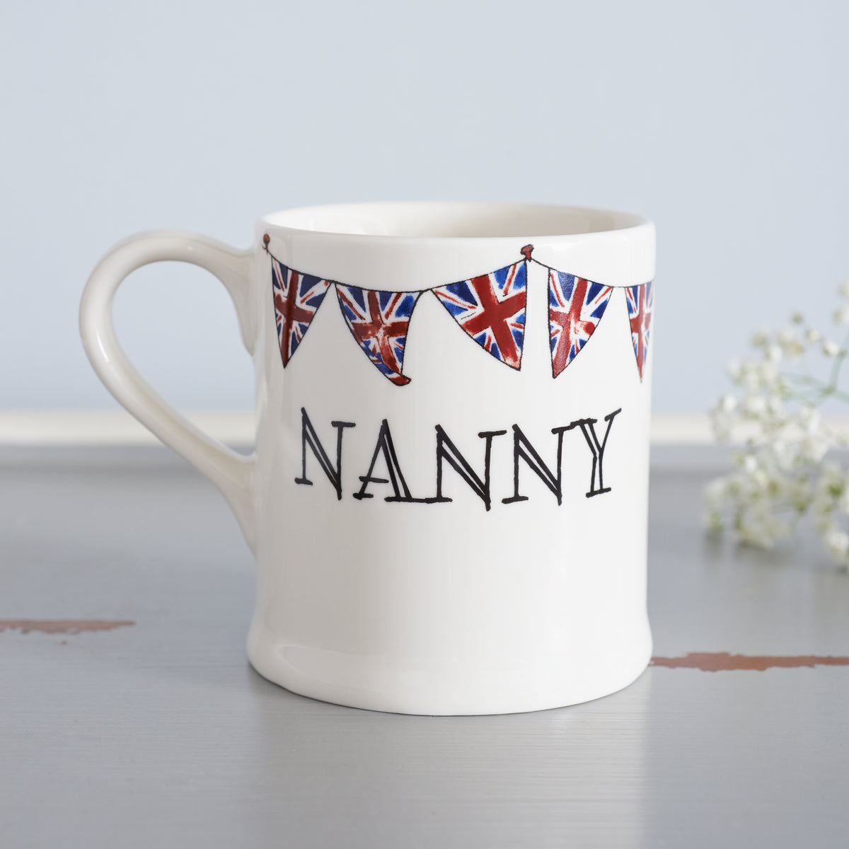 Ceramic Nanny mug with hand-drawn Union Jack bunting, styled beside delicate white flowers, a thoughtful gift for a much-loved grandparent