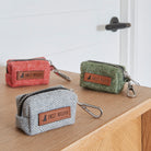 The full Sweet William tweed poo bag holder colour range in grey, green and orange, lined up on a wooden table