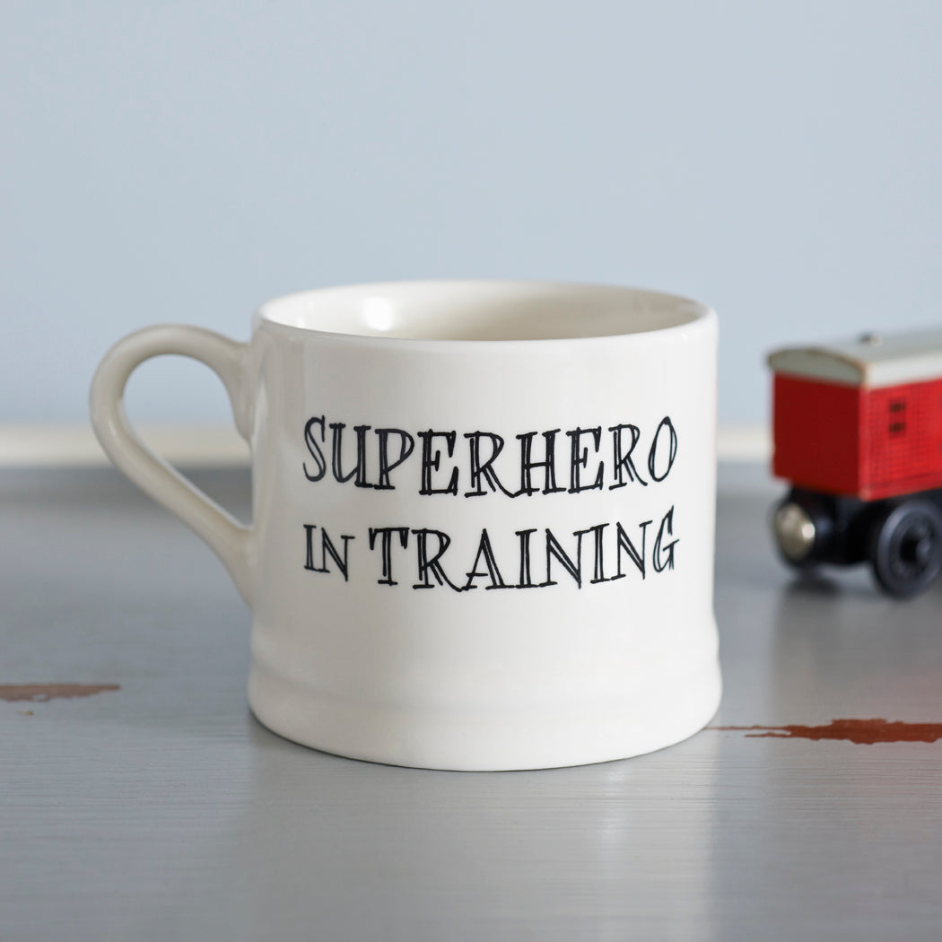 Ceramic “Superhero in Training” baby mug with a red toy train in the background
