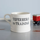 Ceramic “Superhero in Training” baby mug with a red toy train in the background