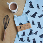 Organic cotton Black Cockapoo / Labradoodle tea towel on a wooden table with a matching mug — organic cotton screen printed kitchen gifts for dog lovers by Sweet William Designs.