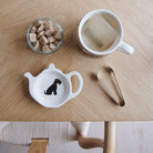 White teapot-shaped Black Schnauzer teabag dish with a black dog illustration and red collar, beside a mug, sugar cubes and sugar tongs