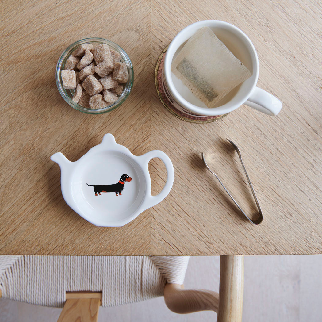 White teapot-shaped Dachshund teabag dish with a black and tan sausage dog illustration and red collar, beside a mug, sugar cubes and sugar tongs