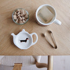 White teapot-shaped Dachshund teabag dish with a black and tan sausage dog illustration and red collar, beside a mug, sugar cubes and sugar tongs
