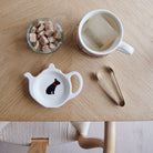 White teapot-shaped French Bulldog teabag dish with a black Frenchie illustration and red collar, beside a mug, sugar cubes and sugar tongs