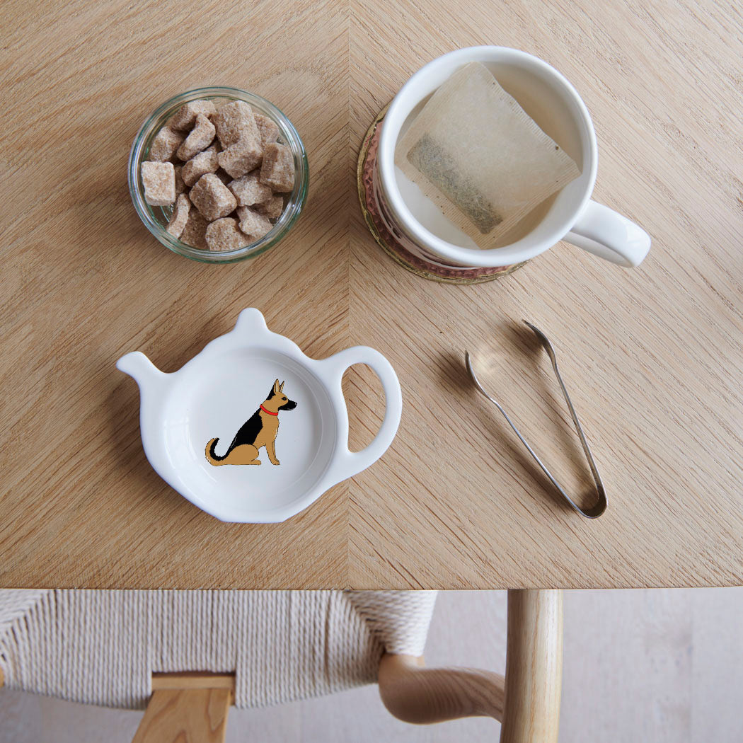 White teapot-shaped German Shepherd teabag dish with a black and tan dog illustration and red collar, beside a mug, sugar cubes and sugar tongs