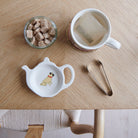 White teapot-shaped Pug teabag dish with a fawn Pug illustration and red collar, beside a mug, sugar cubes and sugar tongs