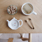 White teapot-shaped Westie teabag dish with a white dog illustration and red collar, beside a mug, sugar cubes and sugar tongs