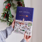 Front of the Christmas Dog Treat Advent Calendar held in a hand, showing a snowy British Christmas village scene with the Mischievous Mutts, by Sweet William Designs