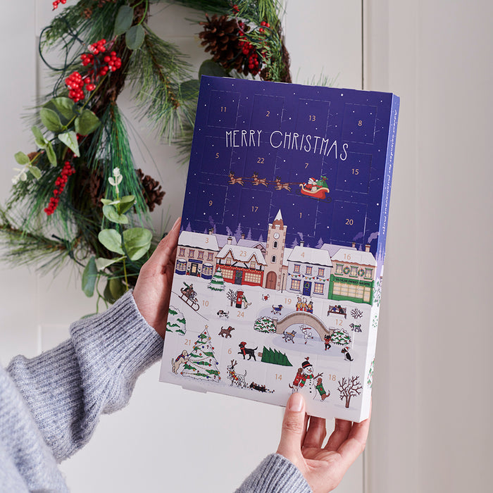 Front of the Christmas Dog Treat Advent Calendar held in a hand, showing a snowy British Christmas village scene with the Mischievous Mutts, by Sweet William Designs