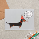 Dachshund 1st birthday card featuring a black and tan sausage dog in a red party hat with a “Happy 1st Birthday!” message, designed by Sweet William Designs