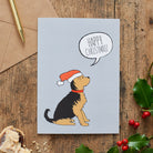 Yorkshire Terrier Christmas card featuring a Yorkie wearing a Santa hat beneath a Happy Christmas message, styled with holly on a wooden table