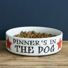 White porcelain “Dinner’s In The Dog” dog bowl with red star accents on a wooden table.