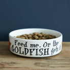 White porcelain cat bowl with “Feed Me Or The Goldfish Gets It” slogan on a wooden table