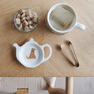 White teapot-shaped Fox Red Lab teabag dish with a fox-red dog illustration and red collar, beside a mug, sugar cubes and sugar tongs